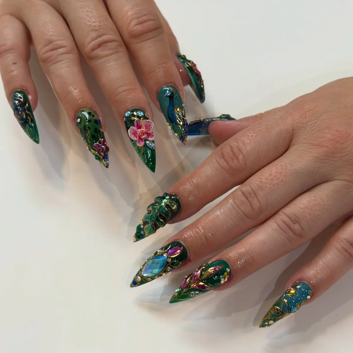 Emerald stiletto nails with a painted peacock, pink orchid, gems and chrome detail.