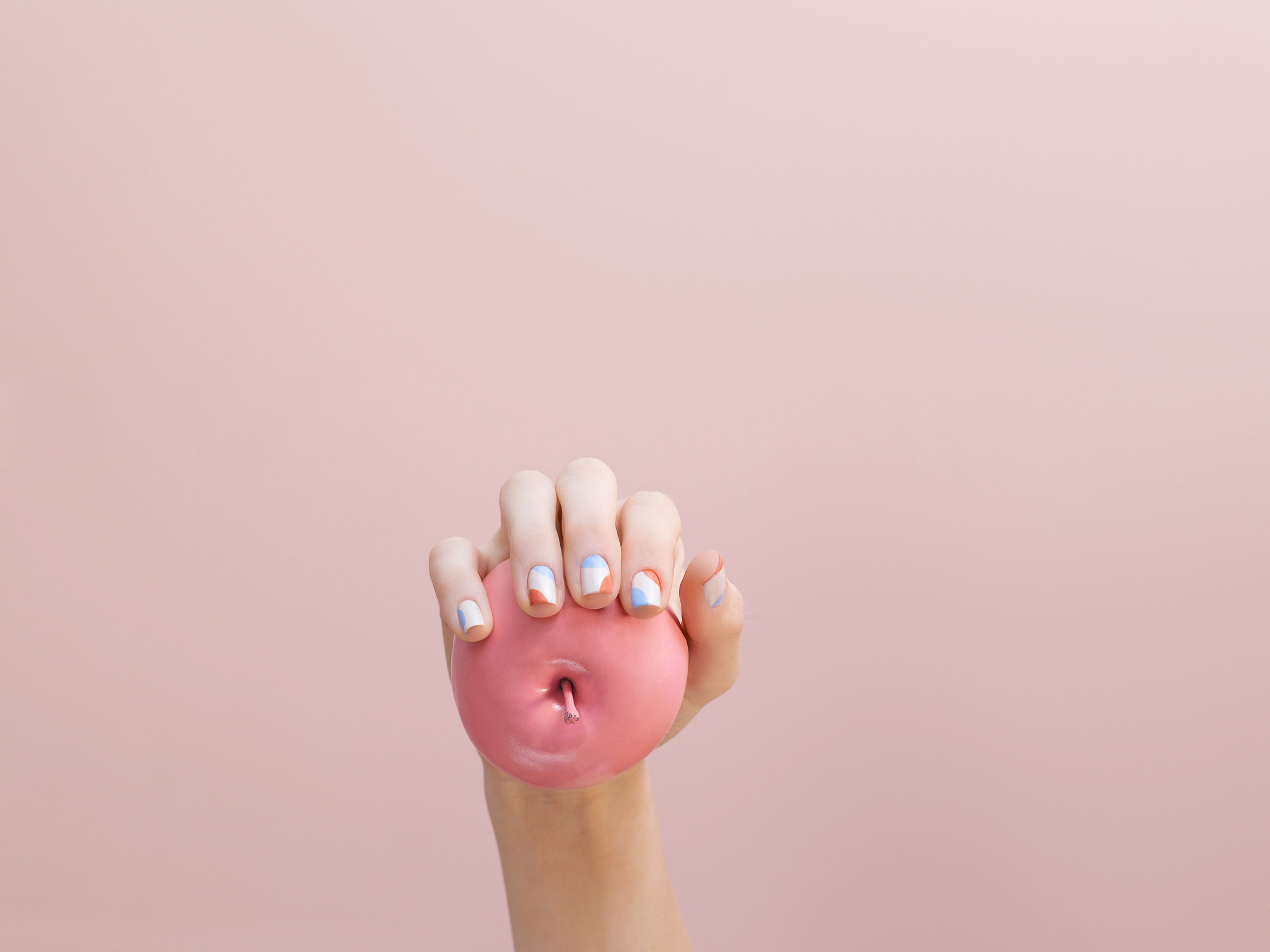 Hand with short square nails in abstract white, blue, and terracotta block nail art holding a matte pink apple against a pink background.