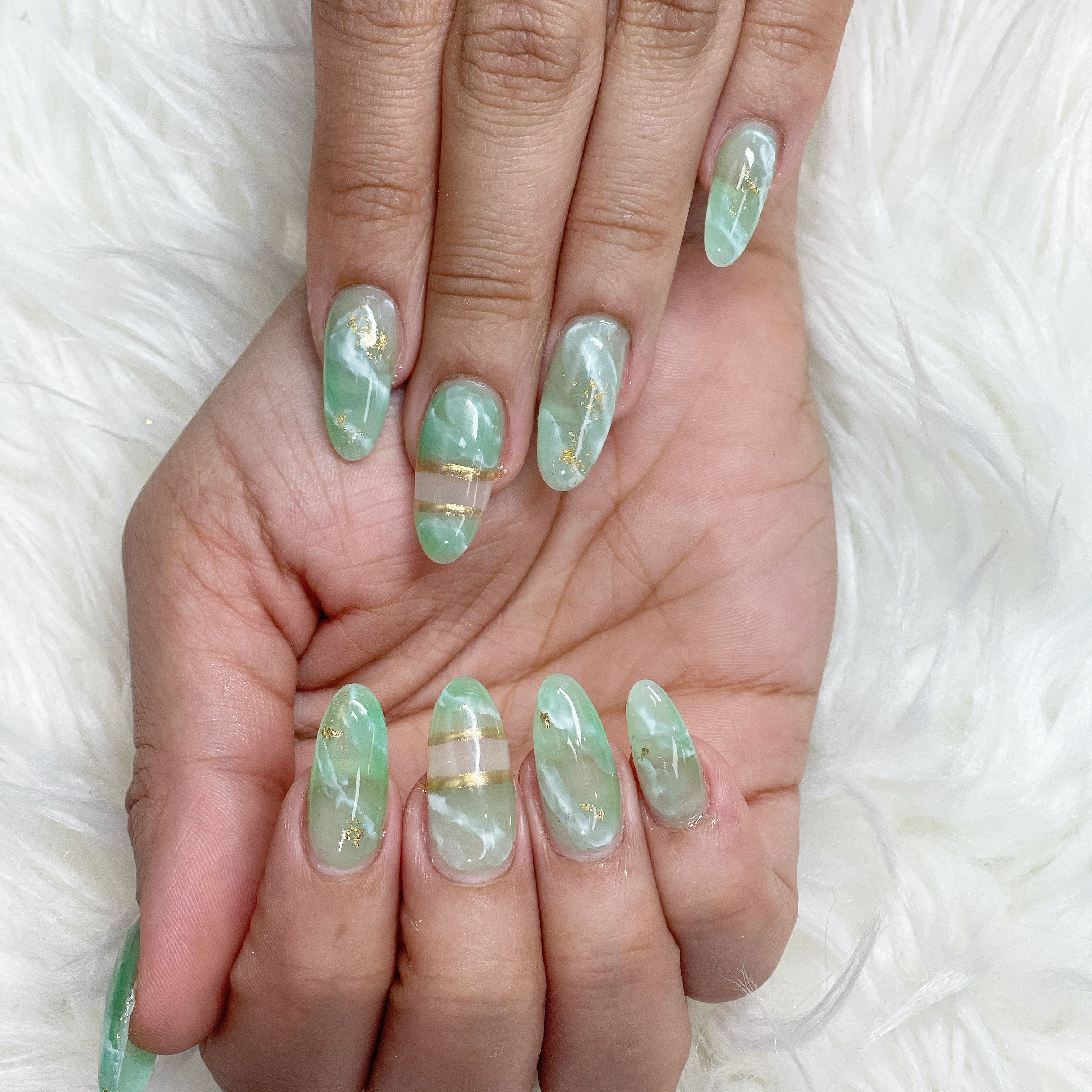 Almond-shaped nails with green and white jade marble, gold foil flakes, and metallic gold horizontal stripe accents on white faux fur.