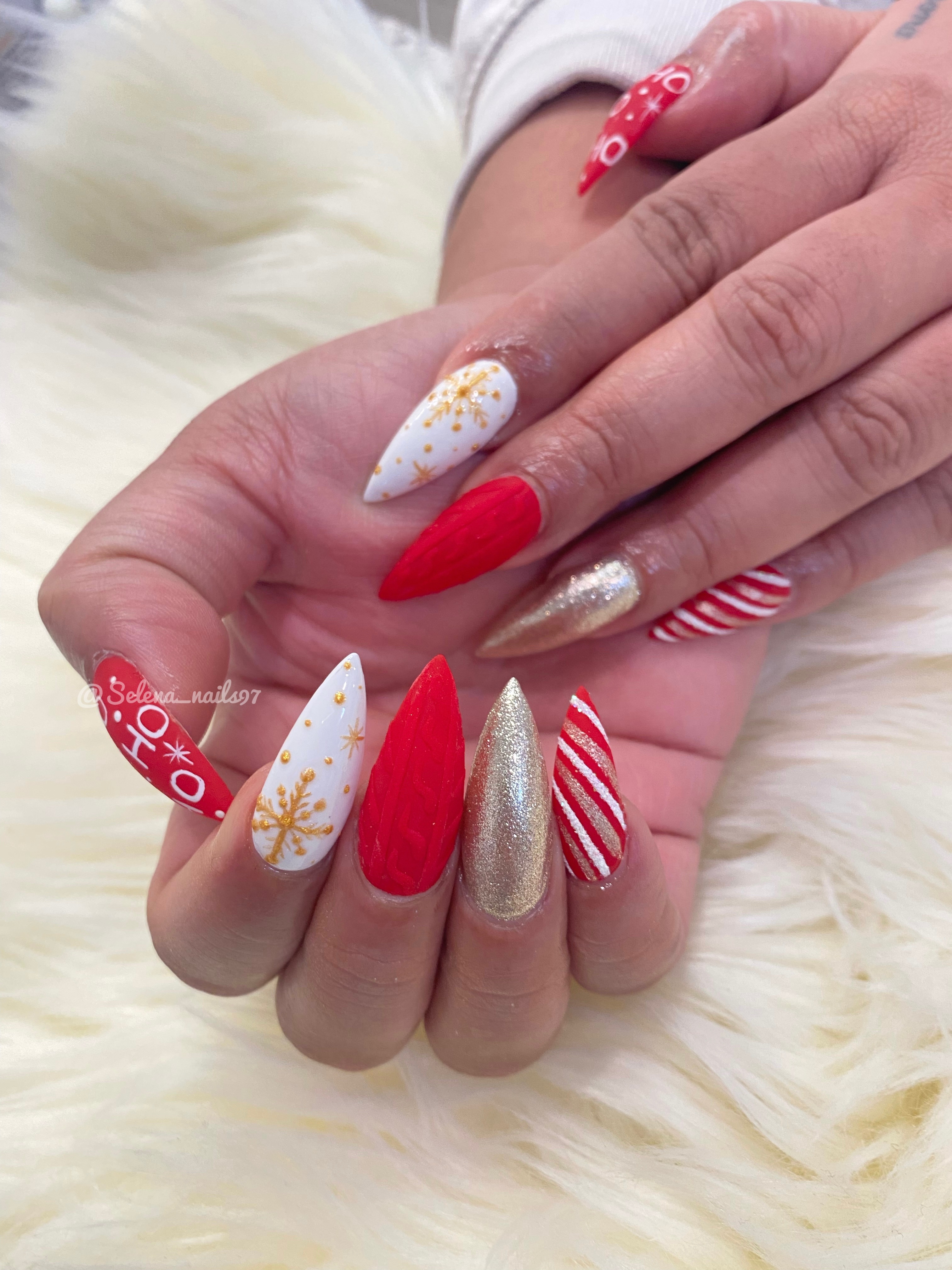 Long stiletto holiday nails with red, white candy-cane stripes, gold glitter, snowflakes, and HO HO HO accents on white faux fur.