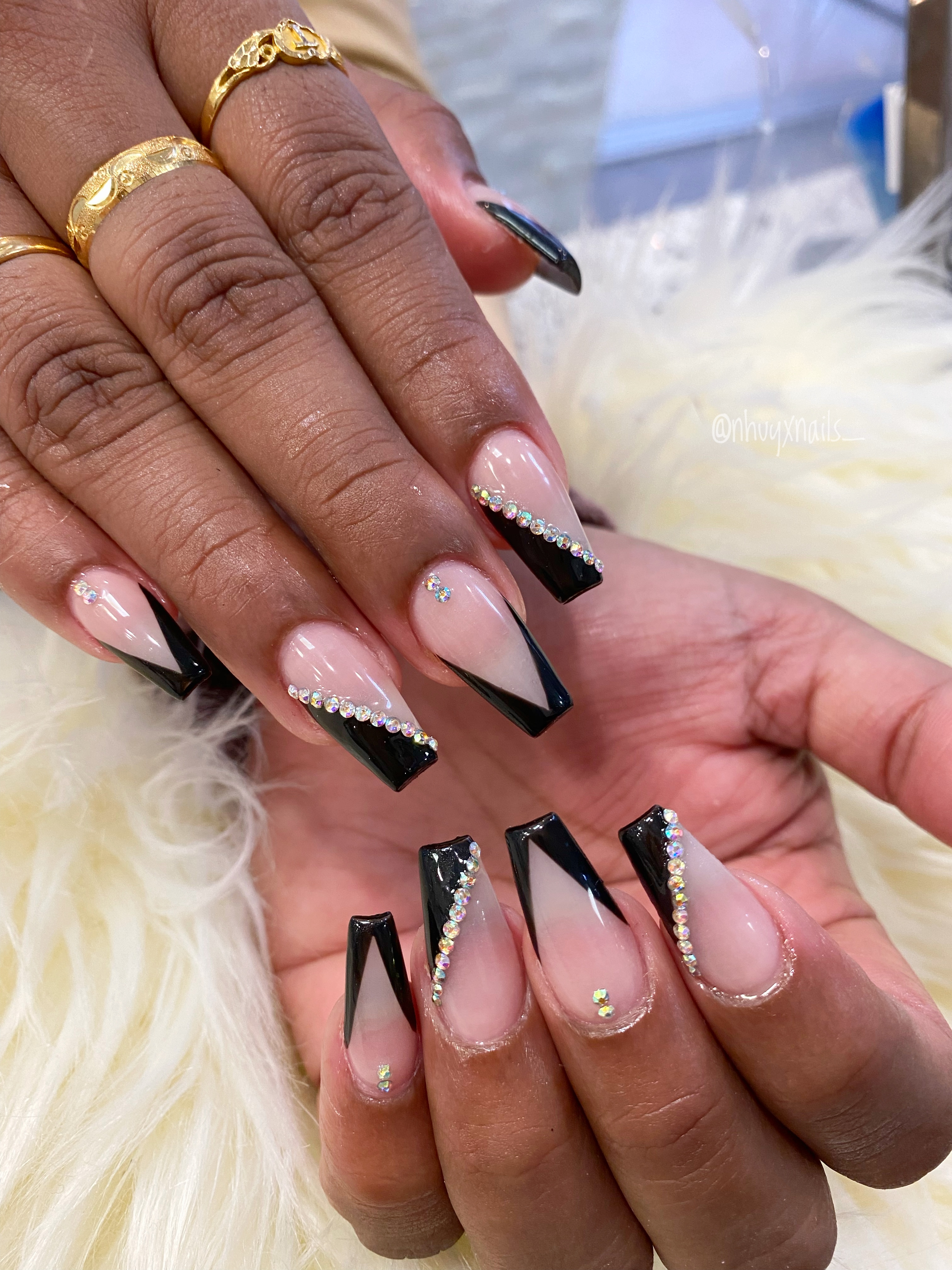 Long coffin-shaped nails with a nude base, black geometric French tips, and small rhinestones, worn with gold rings on white faux fur.