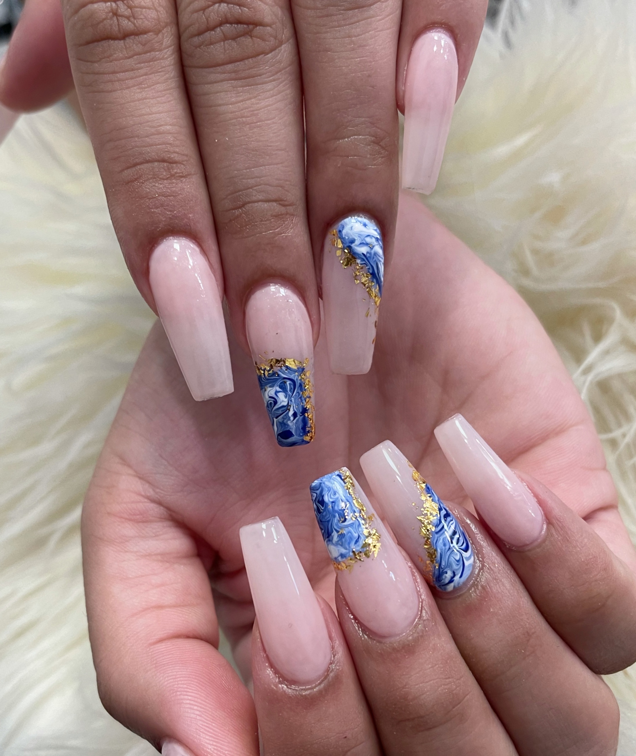 Long coffin-shaped nails with a glossy nude base and blue-and-white marble accent designs bordered by gold foil flakes on white faux fur.