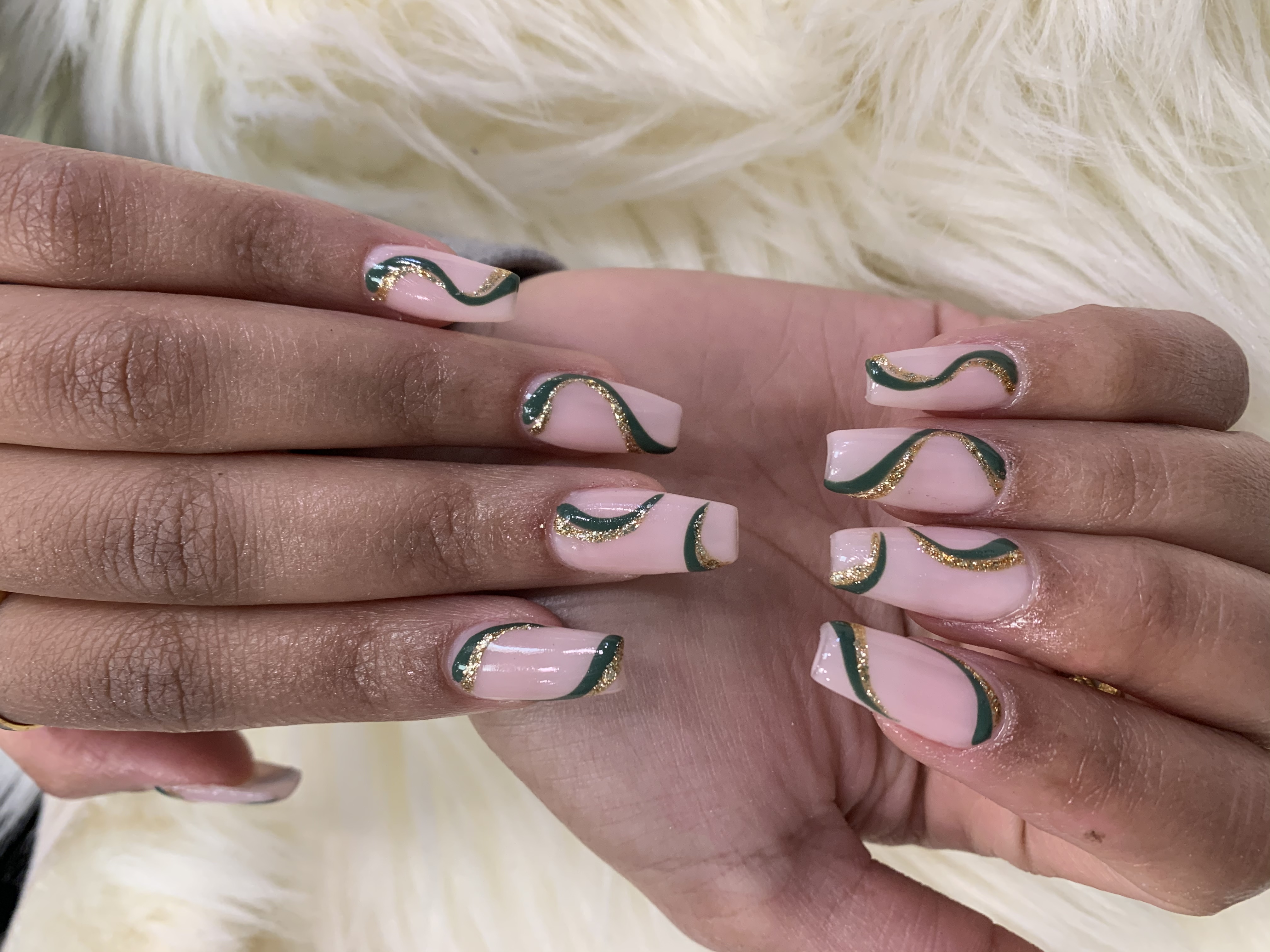 Long coffin nails with a pale nude base, forest-green abstract swirls, and gold glitter outlines on white faux fur.