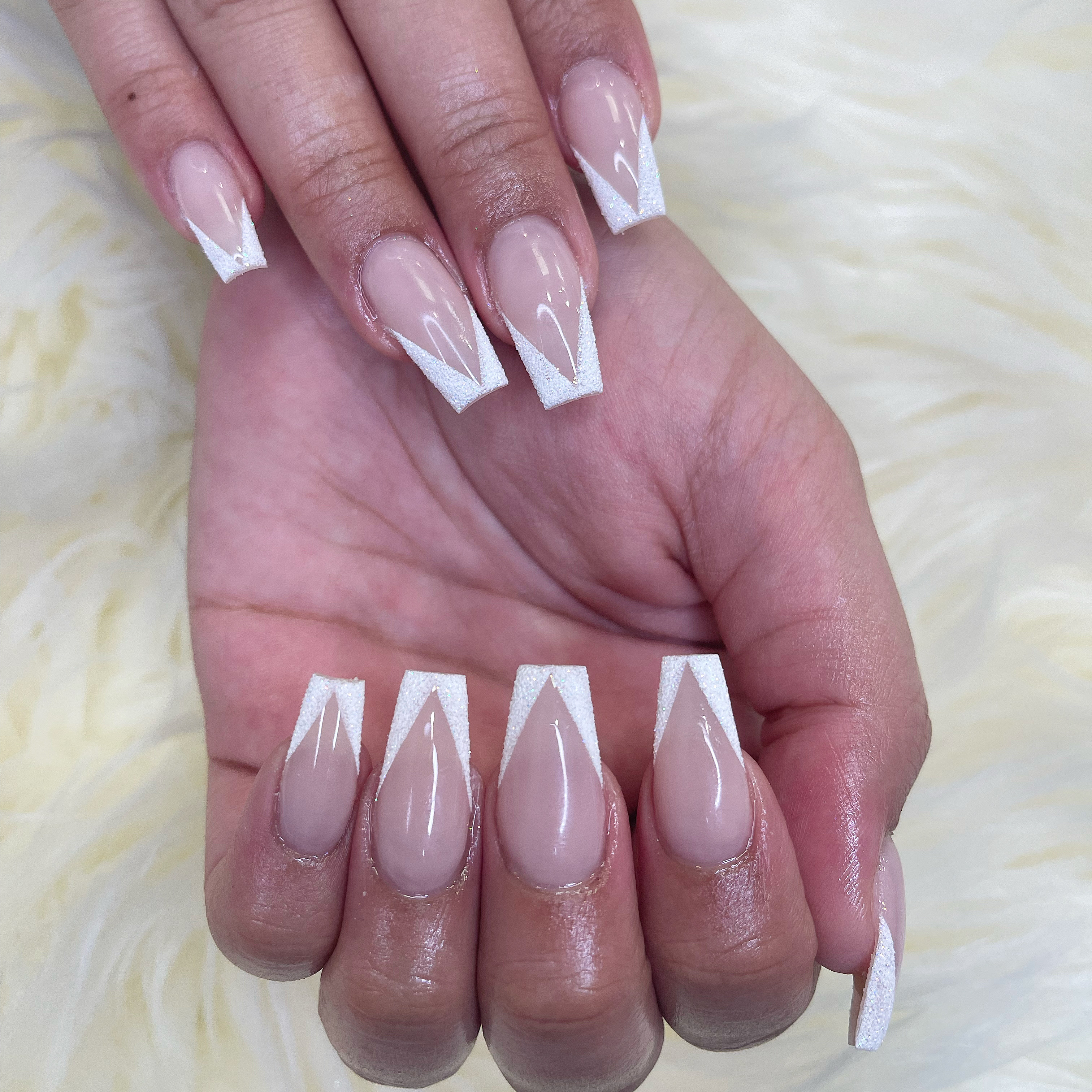 Long coffin nails with a sheer nude base and textured sparkling white V-shaped French tips on a white shaggy background.