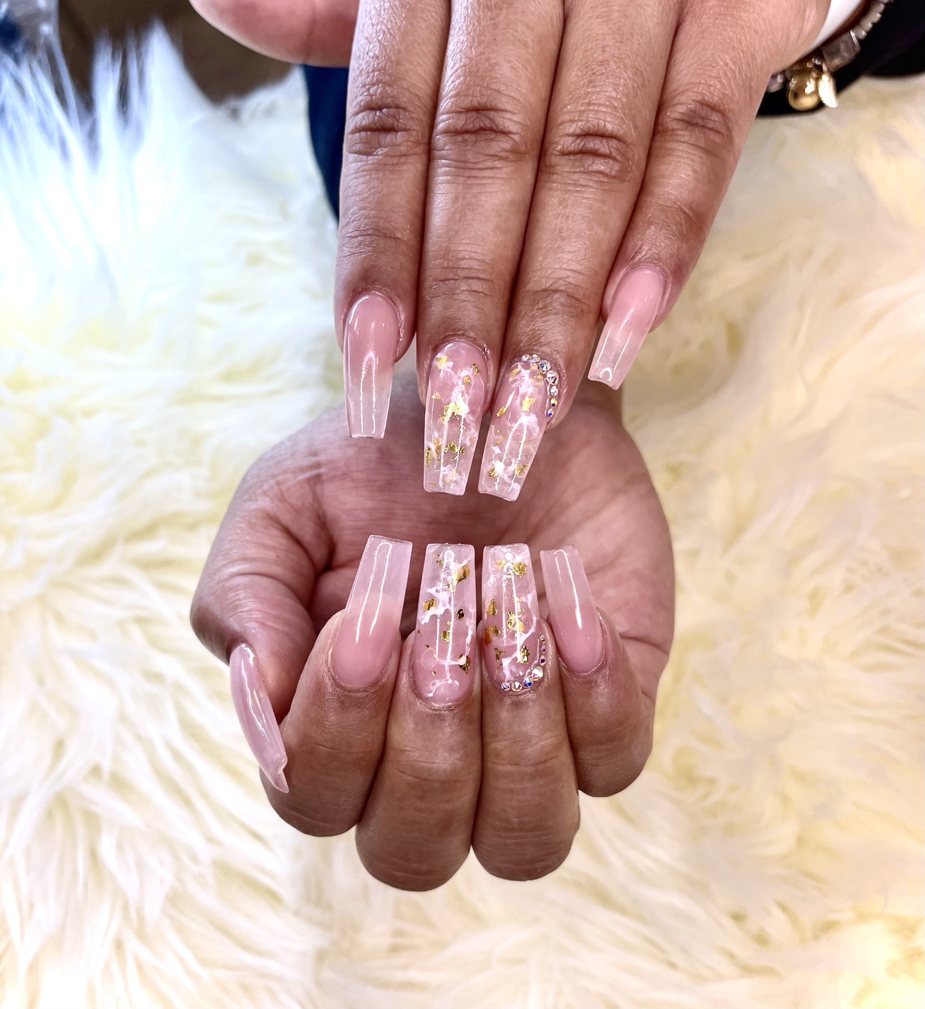 Long coffin nails mixing translucent pink polish with pink-and-white marble, gold foil flakes, and rhinestone accents on white faux fur.
