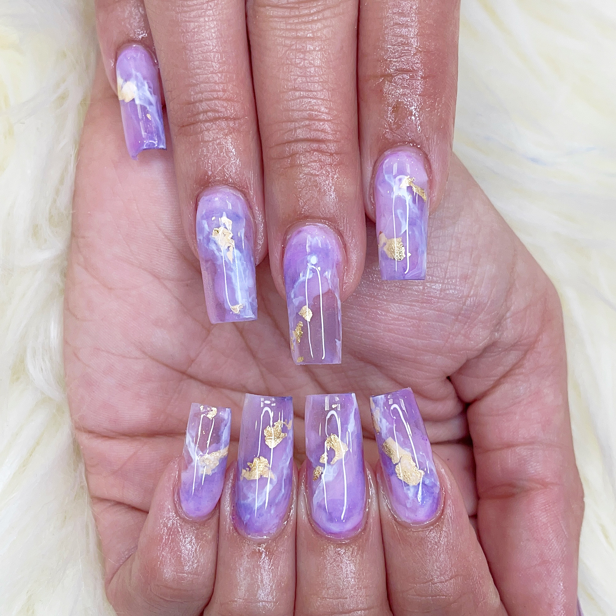 Long square nails with lavender and white marble swirls and scattered gold foil flakes on a white faux-fur background.