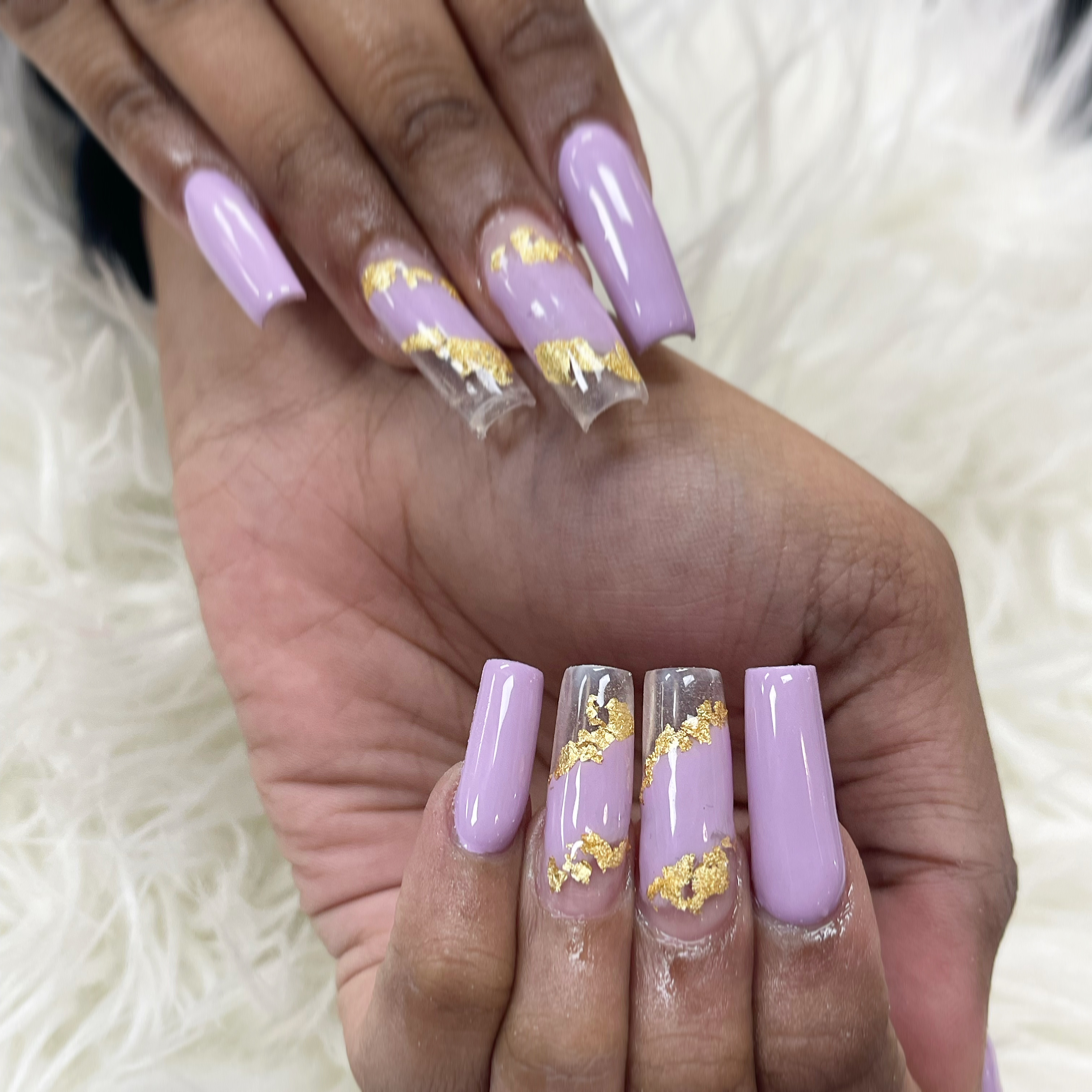 Long square nails in light lavender with clear tips and diagonal purple bands edged in jagged gold foil on white faux fur.
