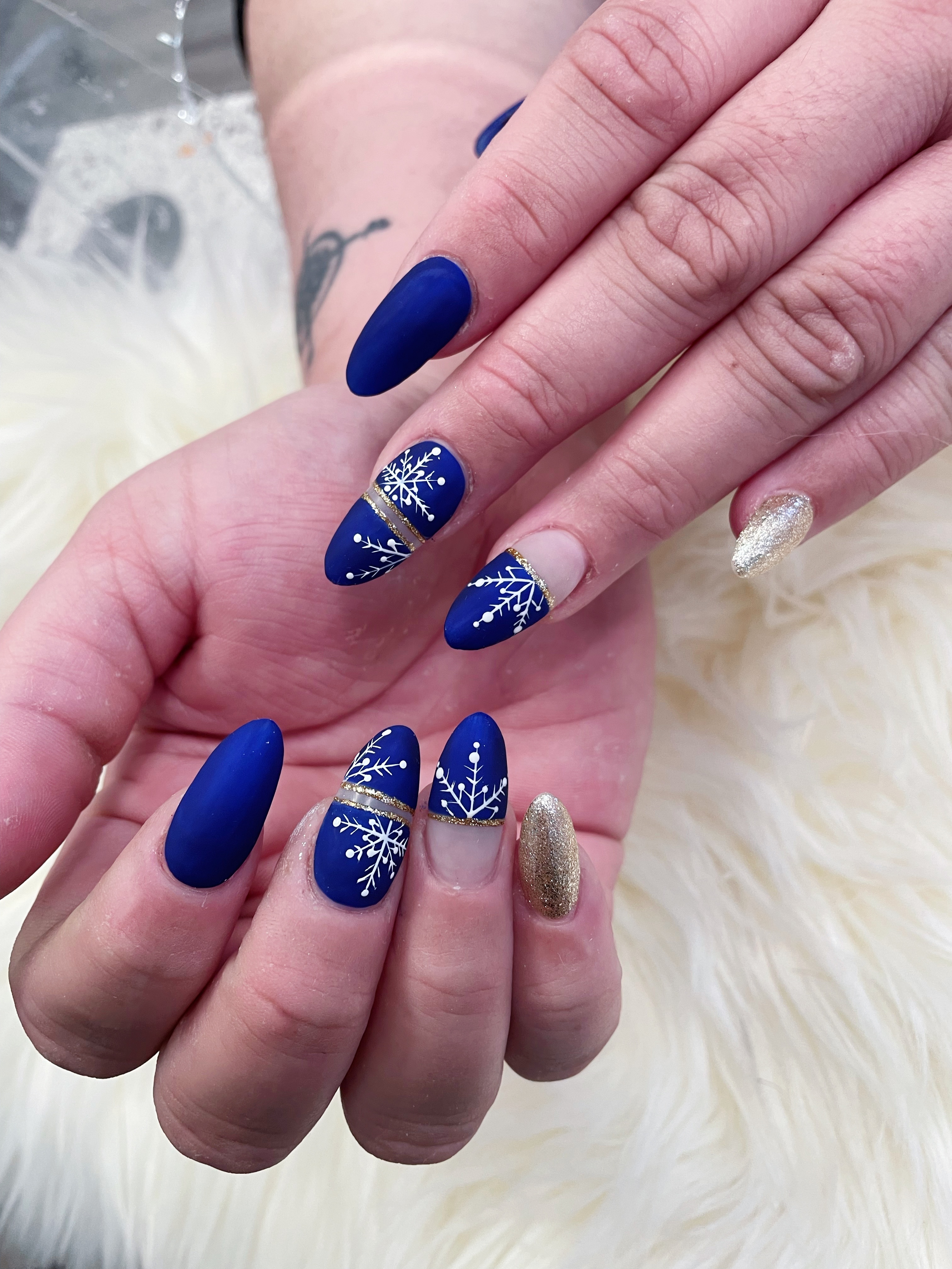 Almond nails in matte royal blue with white snowflake art, negative-space gold glitter stripes, and full gold glitter pinkies on white faux fur.