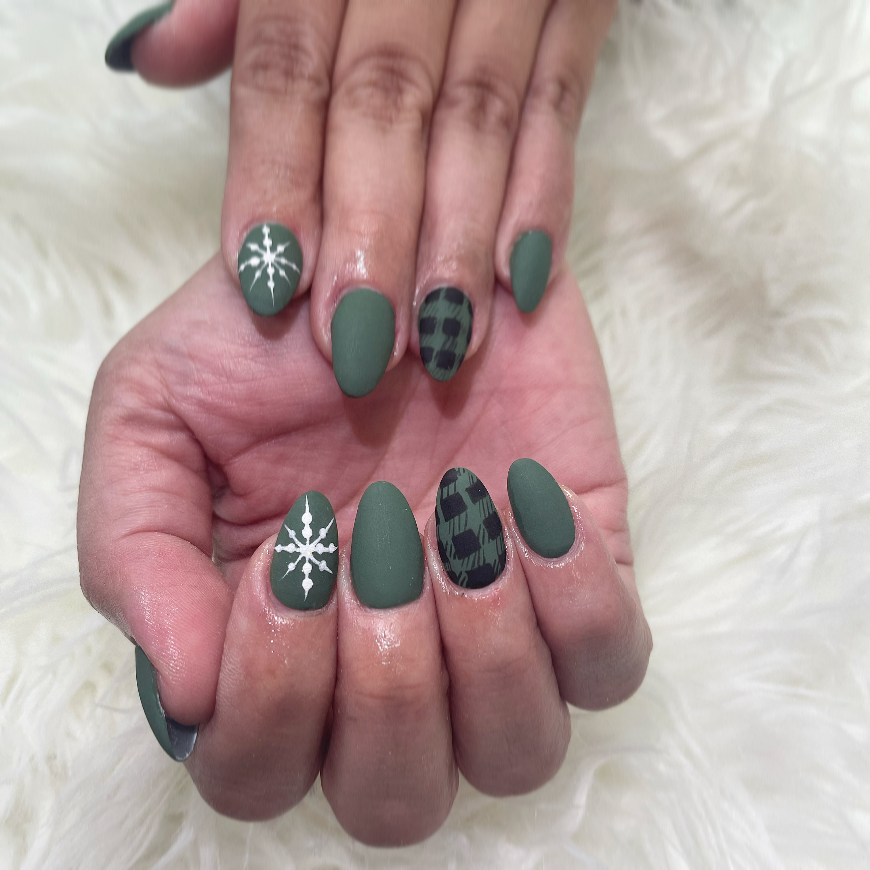Almond-shaped matte forest-green nails with white snowflake art and dark green buffalo-plaid accent nails on white faux fur.