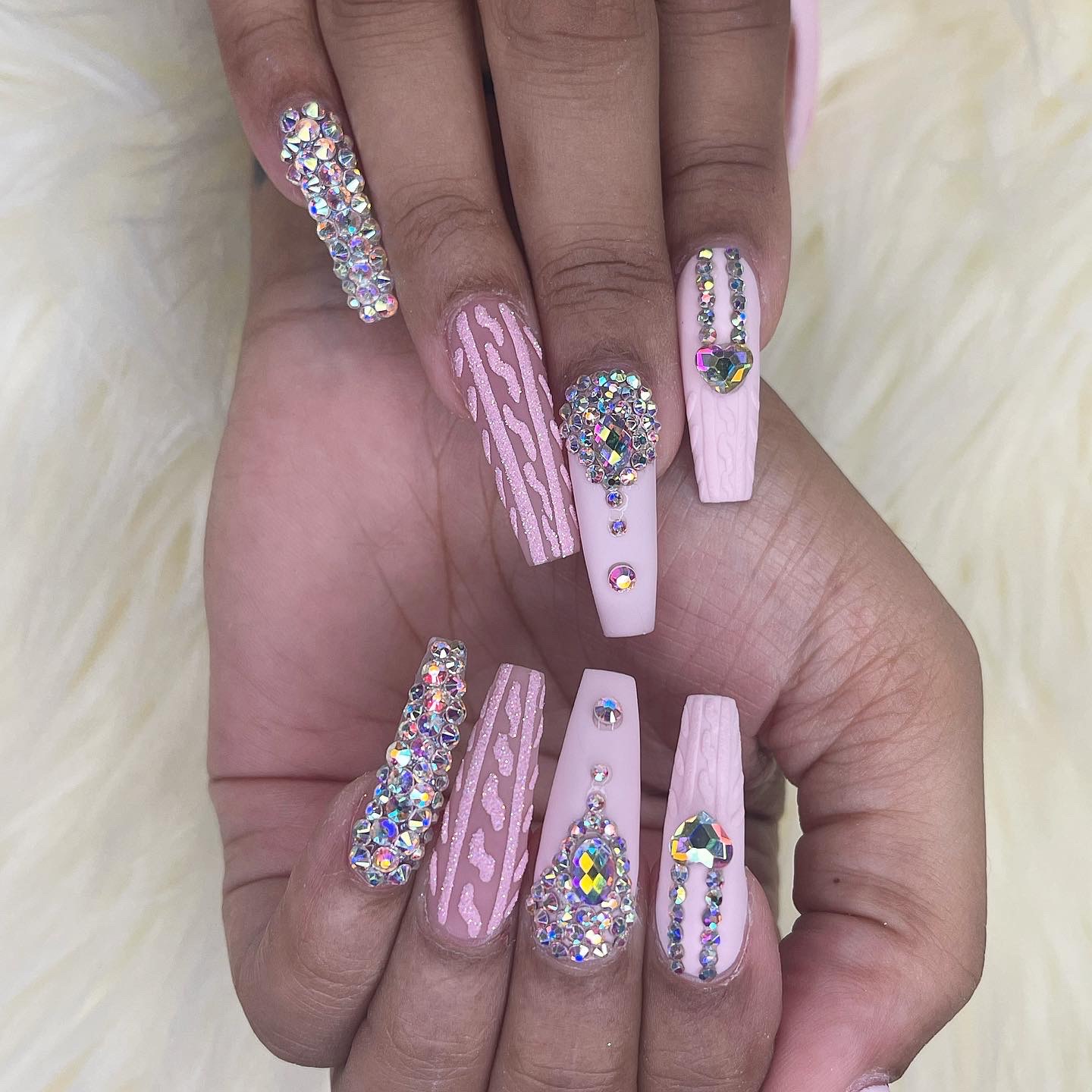 Long matte pink coffin nails with 3D cable-knit texture and heavy iridescent rhinestone embellishments on white faux fur.