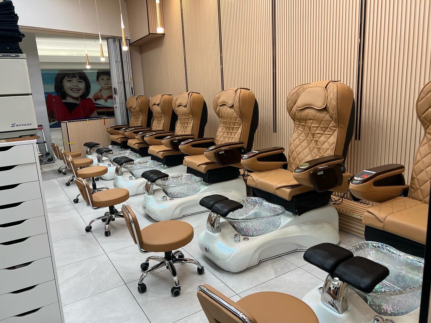 Wide view of a row of tan pedicure massage chairs with white basins and matching technician stools in a clean salon with wood-paneled walls.
