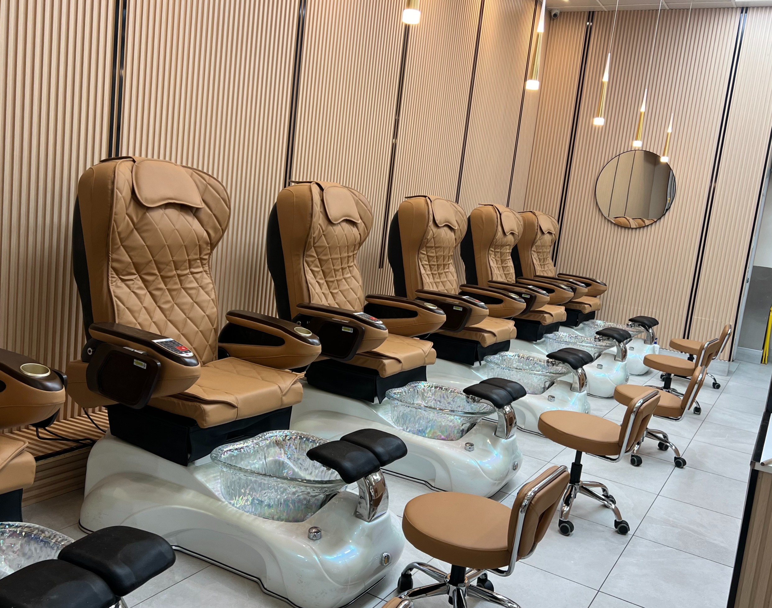 Row of tan leather pedicure spa chairs with clear glass foot basins against a light wood-slatted wall in a modern nail salon.