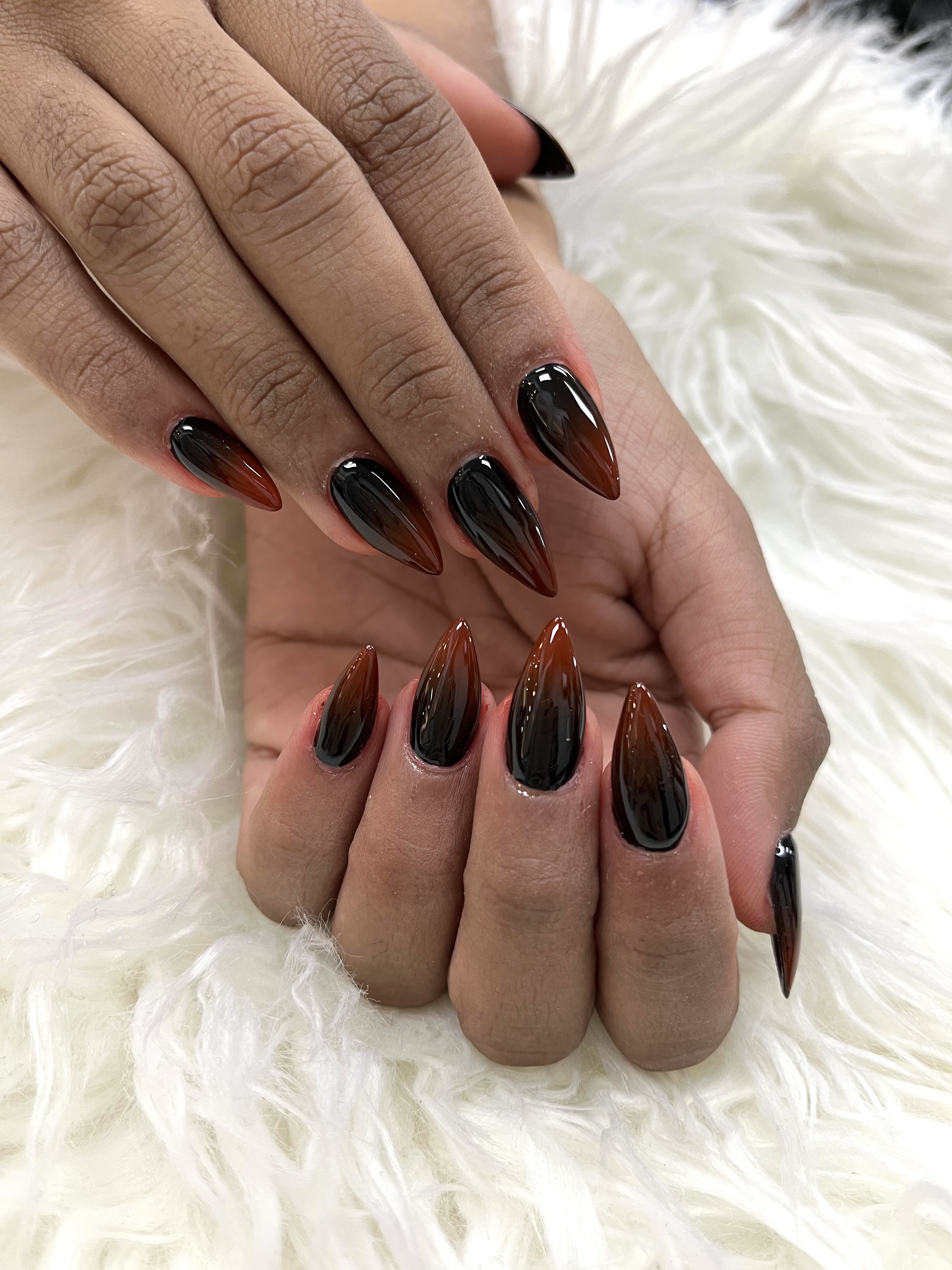Long stiletto nails with a glossy black-to-amber ombre gradient resting on white faux fur.